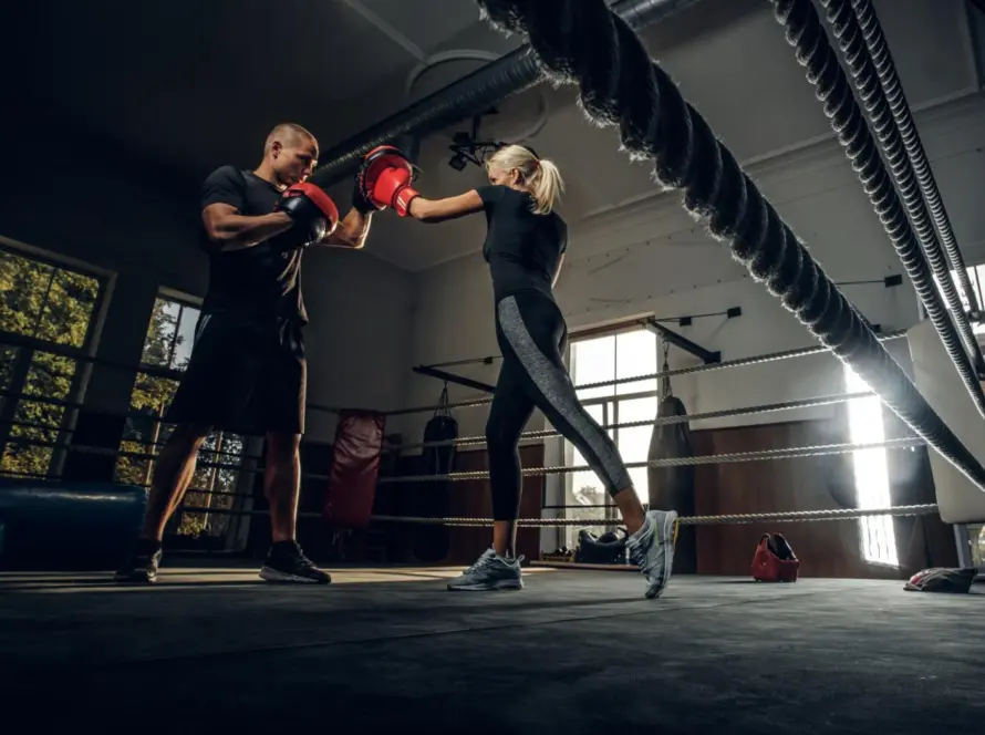 Entrenador y boxeadora practicando técnicas de boxeo en un gimnasio, con guantes rojos y un ambiente iluminado.
