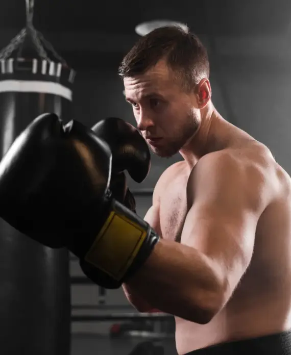 Boxeador en un gimnasio, con guantes de boxeo negros, concentrado en su técnica frente a un saco de boxeo. Fondo con iluminación suave y ambiente de entrenamiento.