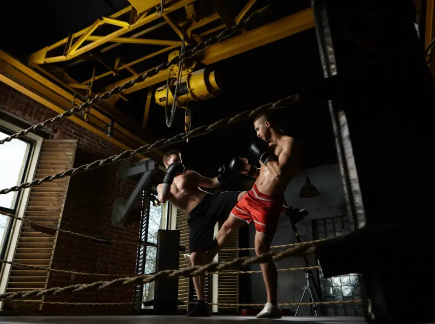 Dos boxeadores entrenando en un ring, uno lanza una patada mientras el otro se prepara para bloquear. El ambiente es industrial, con paredes de ladrillo y luces tenues.
