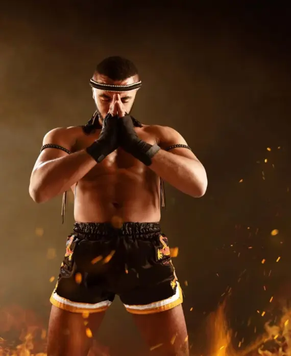 Hombre musculoso en postura de meditación, vestido con shorts de combate, rodeado de llamas y chispas, en un fondo dramático que evoca fuerza y concentración en artes marciales.