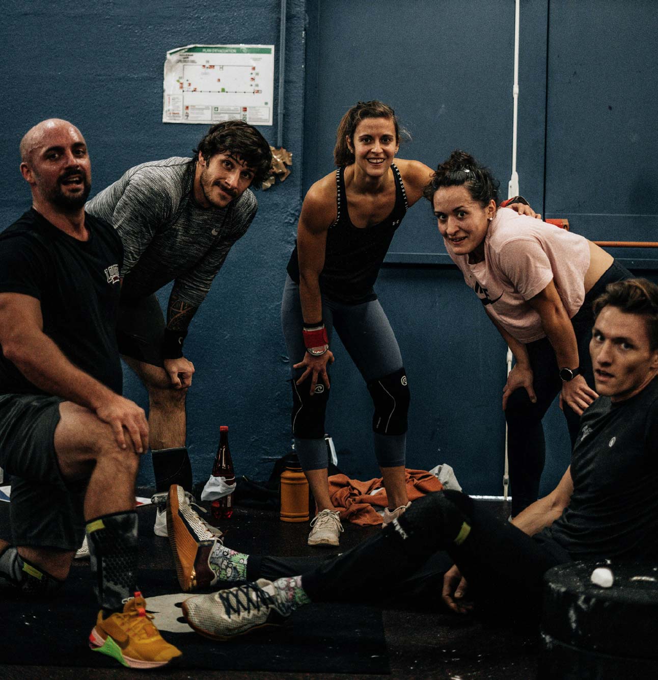 Grupo de cinco personas en un gimnasio, posando después de un entrenamiento intenso. Dos hombres y tres mujeres muestran sonrisas y un ambiente de camaradería. Se pueden ver botellas de agua y equipo de entrenamiento en el fondo, lo que sugiere un enfoque en fitness y bienestar.