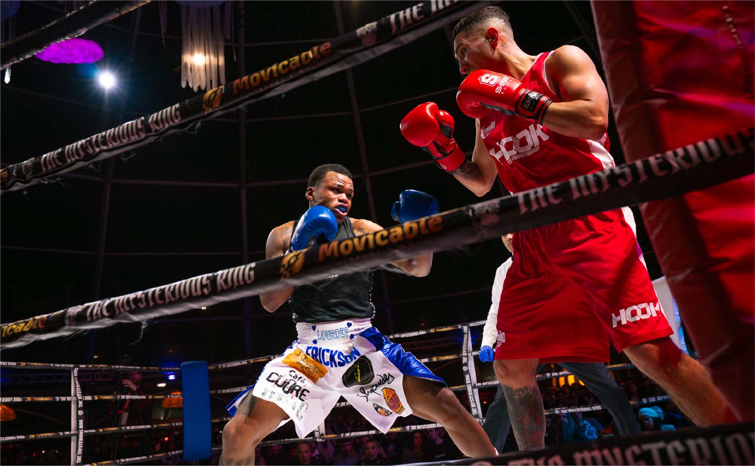 Boxeadores en acción en el ring, uno con guantes azules y el otro con guantes rojos, durante una pelea emocionante. El ambiente está iluminado y lleno de espectadores.