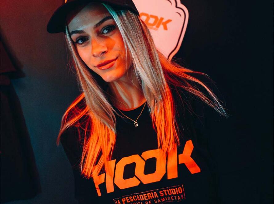 Mujer con camiseta negra y gorra de la marca "HOOK", posando frente a un fondo oscuro con iluminación roja, representando un ambiente moderno y urbano. La camiseta destaca el nombre "HOOK" y el lema "La Pescadería Studio, la fábrica de camisetas".