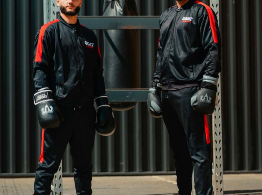 Dos boxeadores de pie frente a un saco de boxeo, vistiendo trajes deportivos negros con detalles rojos y guantes de boxeo, en un entorno de entrenamiento al aire libre.