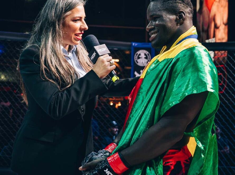 Entrevista a un luchador de MMA en el octágono, con una presentadora sosteniendo un micrófono. El luchador lleva una capa con los colores de su bandera, mientras sonríe durante la conversación. Fondo del evento deportivo con espectadores y pantallas mostrando otros luchadores.