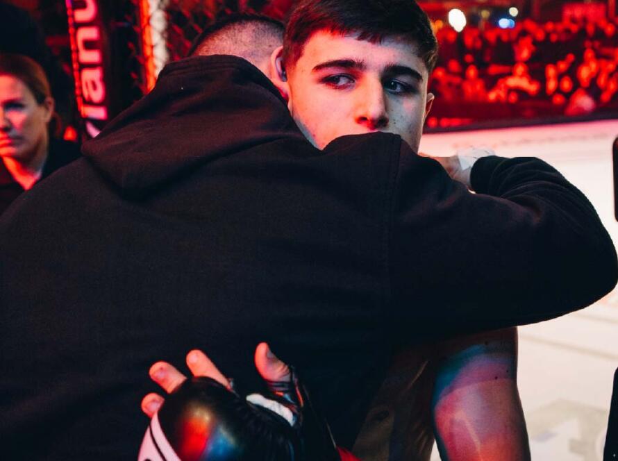 Luchador de MMA recibiendo apoyo emocional antes de una pelea, con guantes de boxeo y un entorno iluminado por luces de colores en un evento deportivo.