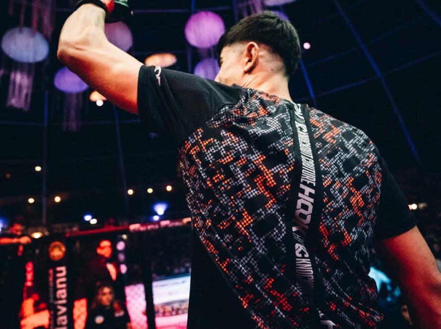 Luchador levantando el puño en señal de victoria, vistiendo una camiseta de diseño moderno, con una jaula de combate y espectadores al fondo, en un evento de artes marciales mixtas.