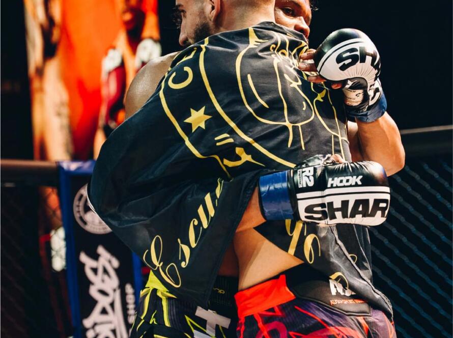 Dos luchadores de MMA se abrazan en el octágono, mostrando camaradería y respeto después de una pelea intensa. Ambos llevan guantes y shorts de combate con diseños llamativos, mientras que el fondo presenta una pantalla con imágenes relacionadas con el evento.