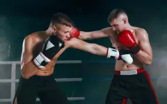 Dos boxeadores en un ring, uno con guantes negros y otro con guantes rojos, intercambiando golpes. La imagen muestra la intensidad del combate en un ambiente de gimnasio.