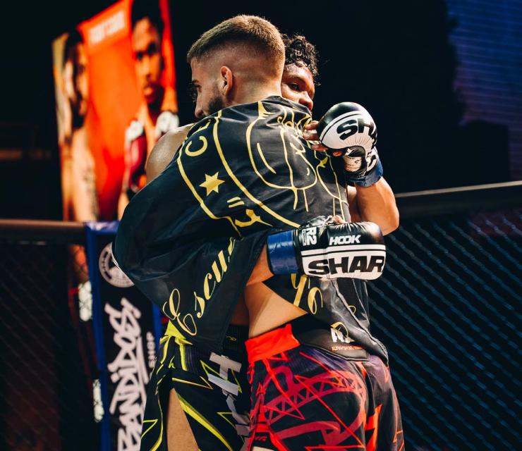 Dos luchadores se abrazan en el ring tras una pelea, mostrando camaradería y respeto. Uno lleva una toalla negra con diseño dorado y el otro usa guantes de boxeo. En el fondo, se ven carteles de otros competidores, destacando la atmósfera de un evento de artes marciales.