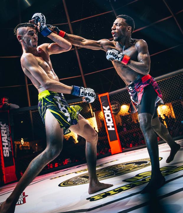 Lucha de artes marciales mixtas en un ring, donde un peleador golpea a su oponente con un puñetazo. Ambos luchadores están en plena acción, mostrando técnicas de combate. El ambiente es intenso, con luces brillantes y una jaula de protección visible.